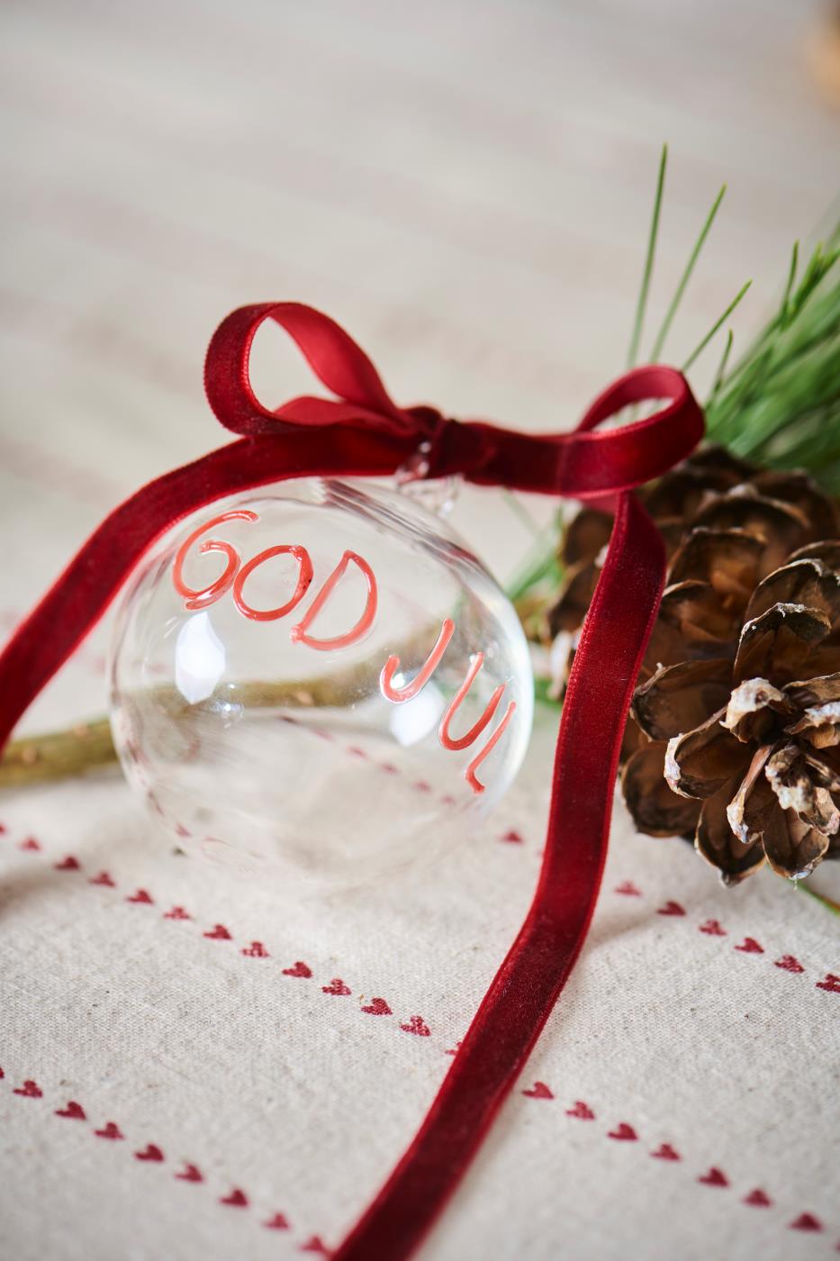 Julekule i klart glass med rød tekst GOD JUL fra Ib Laursen, liggende på et bord med sløyfe