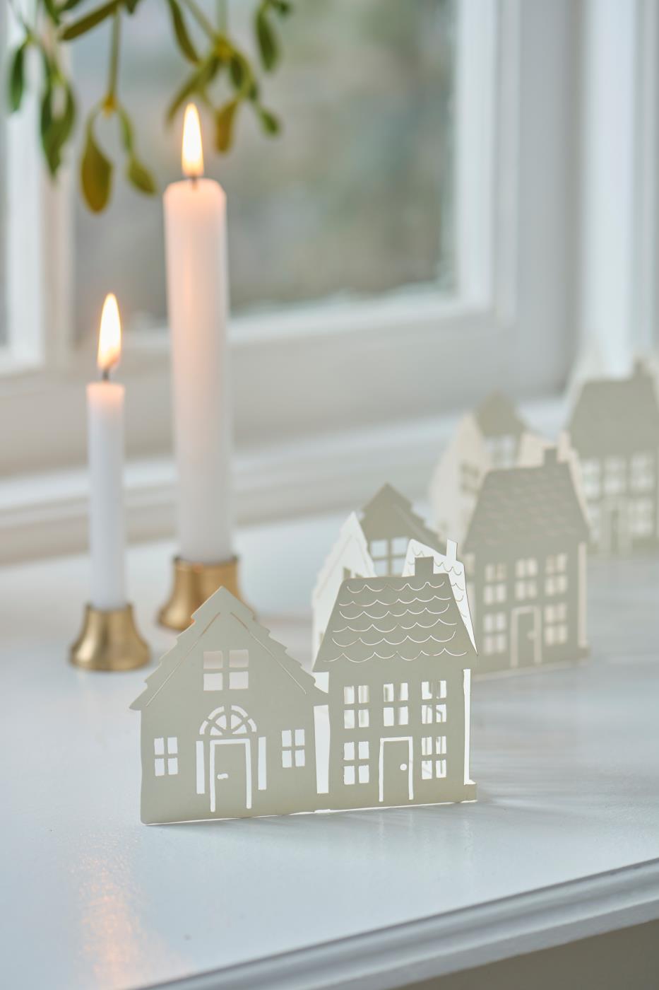 Papirpynt med Hvide huse på række stående i vindueskarm med lys.