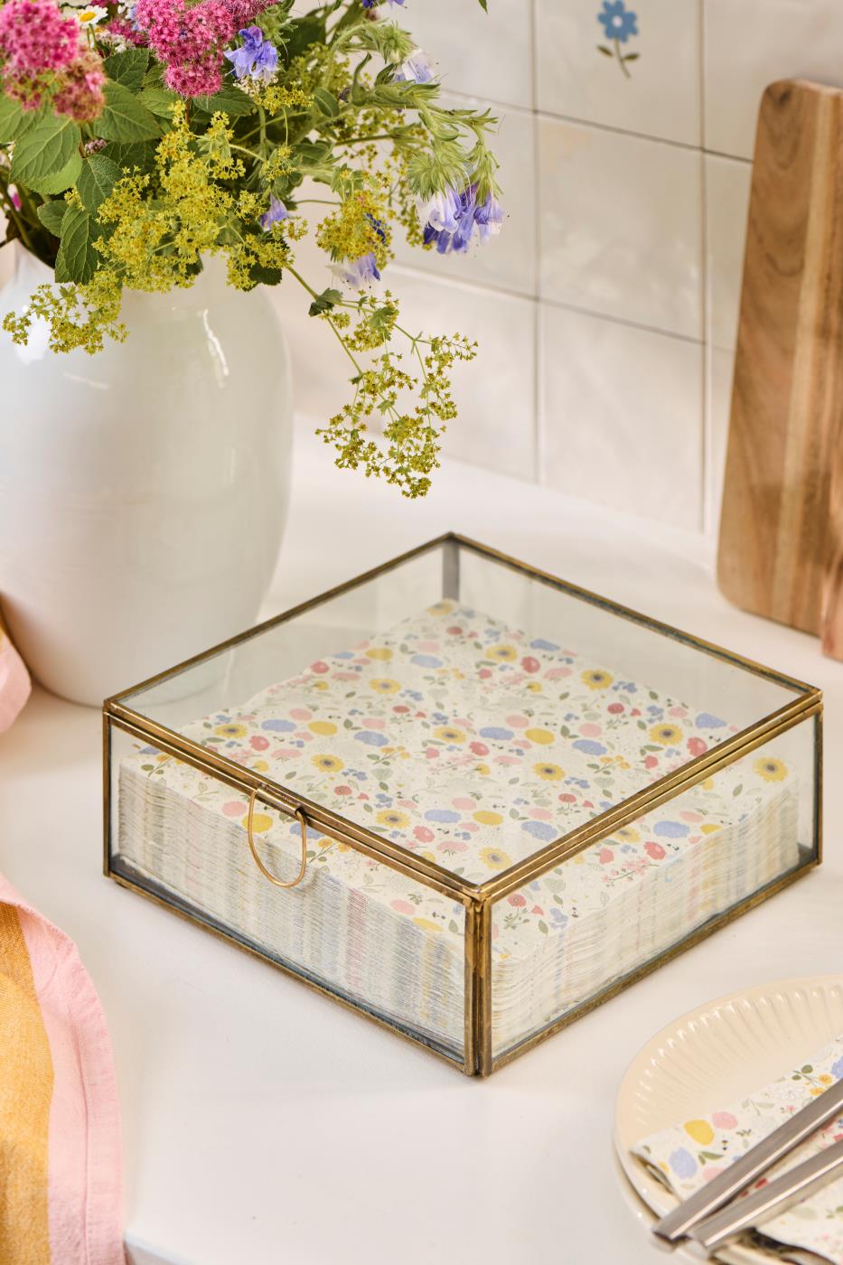 Solitaire glassboks med gullfarget ramme og servietter på et lyst bord ved en vase med blomster